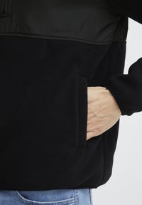 Fekete fleece dzseki sima felső anyaggal. Cipzáras zsebet tartalmaz, és egy kéz látható az oldalsó zsebében, bemutatva a textúrát és az elkészítést.