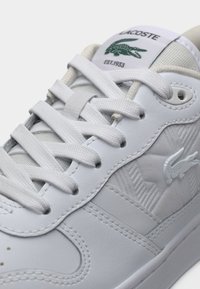 Sneaker en cuir blanc avec côtés texturés, arborant un logo Lacoste vert, lacets plats blancs et languette rembourrée pour plus de confort.