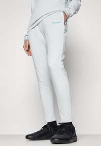 Světle šedé Nike teplákové kalhoty s tapered střihem, s tyrkysovými akcenty, bočními kapsami a elastickým pasem. Nosí se s černými sportovními botami.