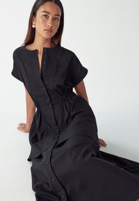 Abito nero con maniche corte, colletto a camicia e vita elastica, con gonna fluente. Tessuto liscio con una leggera trama, design minimalista.