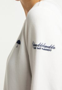 Light grey sweatshirt featuring a navy umbrella graphic and text embroidery on the sleeve. Smooth texture, round neckline, relaxed fit.