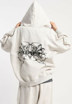 Osoba w jasnoszarej bluzie z kapturem i dresowych spodniach, na której plecach bluzki widnieje czarny abstrakcyjny wzór plemienny.