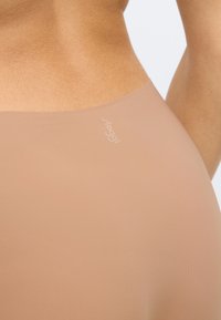 Μπεζ αόρατο εσώρουχο με ομαλή υφή, ελαφρώς καμπυλωτό σχέδιο και μικρό λευκό λογότυπο "sloggi" στη μέση.