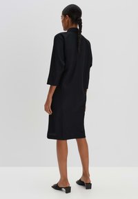 Schwarzes knielanges Kleid mit Dreiviertelärmeln und geradem Schnitt, mit einem Kragen und glatter Textur. Kombiniert mit schwarzen Slippers.