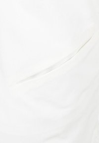 Tela blanca con una textura suave, que presenta un bolsillo lateral discreto con una abertura cosida, mostrando un diseño minimalista.