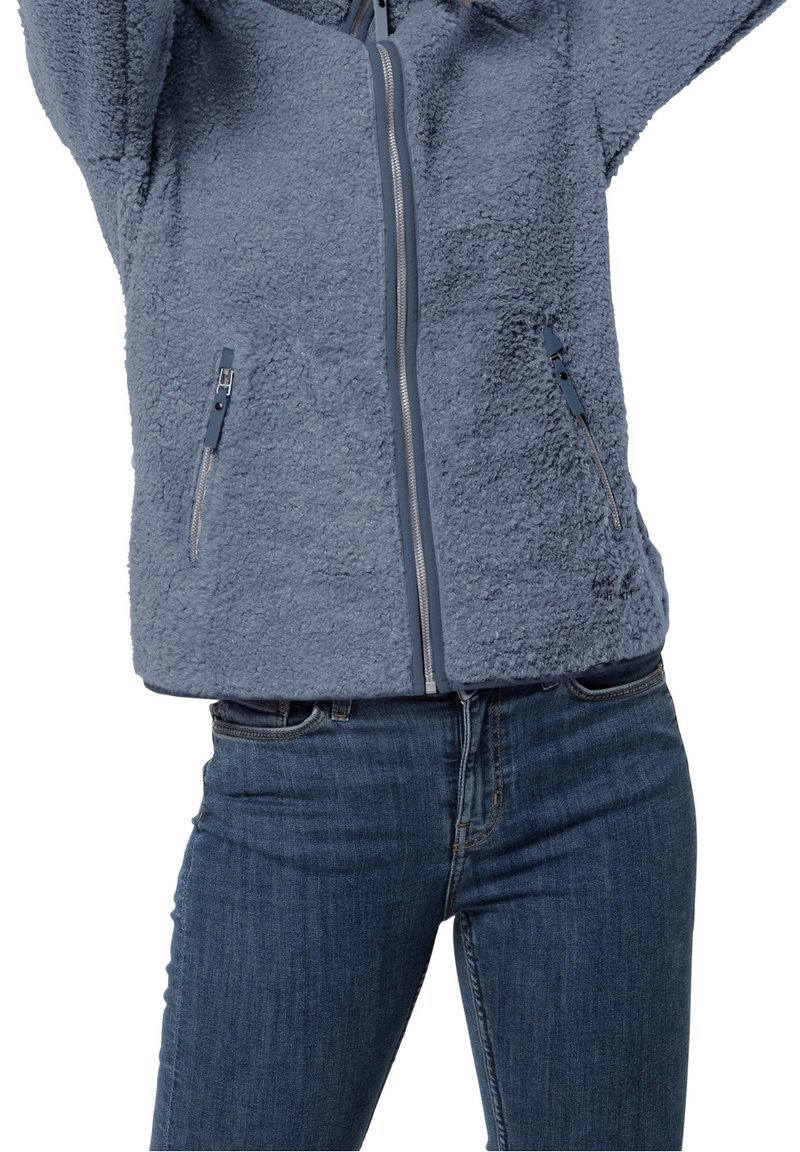 Kurtka z polaru w jasnoniebieskim kolorze z zamkiem błyskawicznym z przodu, dwoma bocznymi kieszeniami i teksturą przypominającą miękkie futro. Połączona z ciemnoniebieskimi dżinsami.