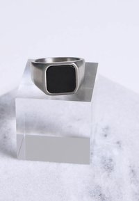 Metalen ring met een vierkante zwarte steen in het midden, geplaatst op een helder acrylblok tegen een lichte marmeren ondergrond. Matte zilveren afwerking.