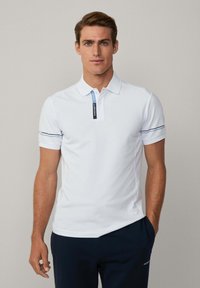 Polo bianca con colletto, strisce blu di accento sulle maniche e un'etichetta di marca sul bottone; texture liscia e design aderente.