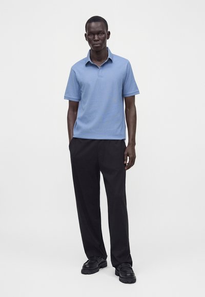 Homme portant un polo bleu clair à manches courtes, pantalon noir et chaussures noires, debout les mains dans les poches sur un fond blanc uni.