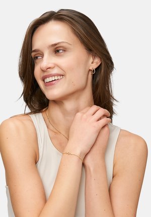 Goldene Creolen und ein zartes Armband mit kleinen Steinen. Das Model trägt ein hellbeiges ärmelloses Oberteil, hat die Haare offen und lächelt.