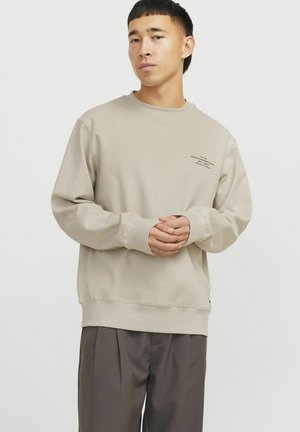 Beige sweatshirt met lange mouwen, ronde hals en geribde manchetten. Heeft subtiele branding op de voorkant gedrukt. Gepaard met bruine broek.