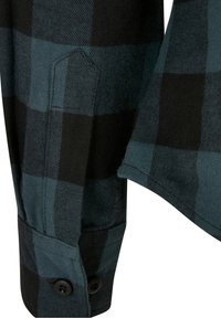 Gros plan sur une manche de chemise à carreaux noirs et vert foncé avec deux boutons noirs sur le poignet et des détails de couture visibles.