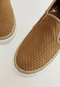 Chaussures slip-on tressées marron avec une surface texturée, bout rond et semelle en caoutchouc blanc. Présente une bordure tressée marron clair autour du bord.