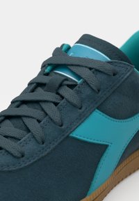 Tamsiai mėlyna zomšinė sportbačių pora su teal akcentais. Ypatumai: tekstūruoti raišteliai, logotipas ant liežuvėlio ir šviesiai ruda gumos padas.
