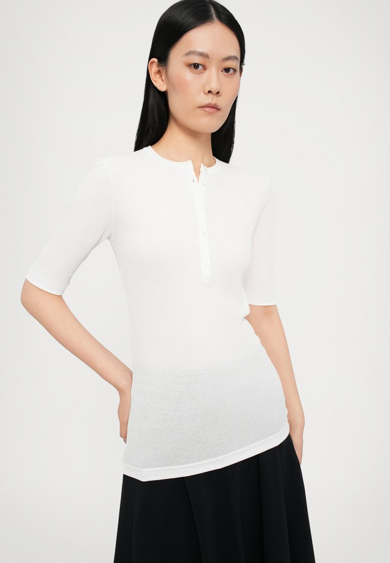 Maglietta henley bianca a maniche corte con scollo rotondo e patta con bottoni, caratterizzata da una texture liscia e leggera e un design leggermente aderente.