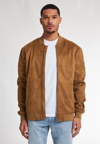 Giacca bomber in finta suede marrone con zip frontale e polsini a coste. Abbinata a una T-shirt bianca e jeans blu. Texture semplice e liscia.