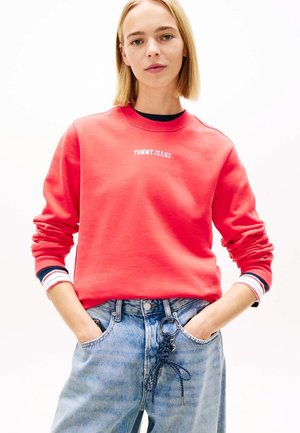 Donna con felpa rossa Tommy Jeans con polsini a righe, jeans a vita alta azzurri e mani nelle tasche su sfondo bianco.