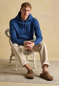 Junger Mann sitzt auf einem Korbstuhl, trägt einen blauen Hoodie, beige Hose und braune Slipper vor einem einfarbigen Hintergrund.