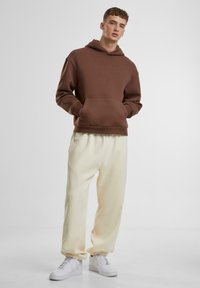 Brun hættejakke med kængurulomme, parret med cremegule sweatpants og hvide sneakers. Blødt stof og afslappet pasform.