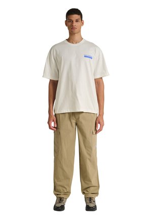 Jonge man die staat, draagt een off-white oversized T-shirt met een klein blauw logo en losse kaki cargobroeken met sneakers.