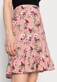 Saia floral rosa com bainha em babados, feita de tecido leve, adornada com flores vermelhas e brancas, e folhas verdes.