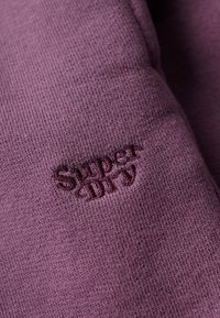 Nærbillede af lilla strikket stof med mørkelilla broderet "Super Dry"-logo centreret på materialet.