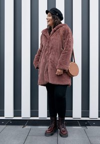 Dirbtinio kailio paltas dulkėto rožinės spalvos, per didelis, su dideliu apykakle, dėvimas ant juodų kelnių. Papuoštas apvalia ruda rankine ir juodais batais.