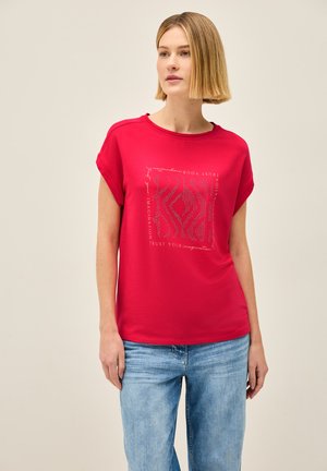 T-shirt rouge à manches courtes avec un col rond, présentant un design graphique en texte blanc et argent, associé à un jean en denim bleu clair.