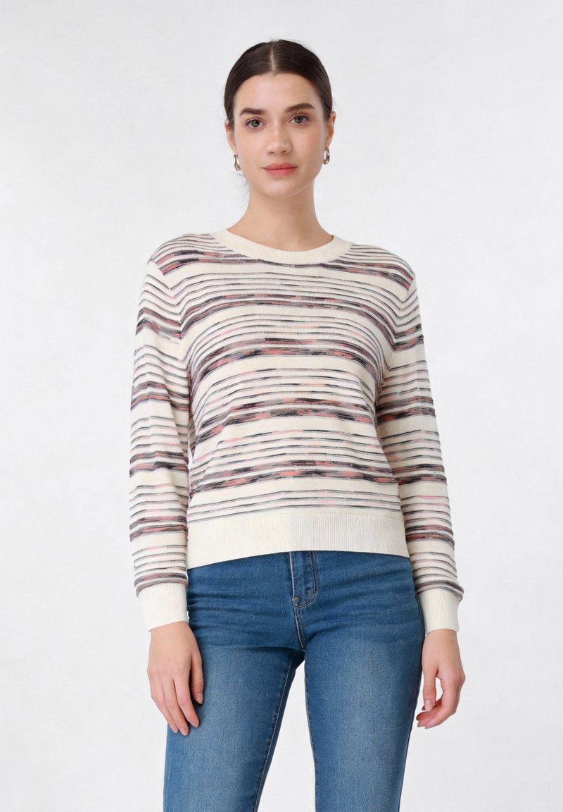 Femme aux cheveux foncés portant un pull rayé blanc, noir et rose ainsi qu'un jean bleu, debout devant un fond clair uni.