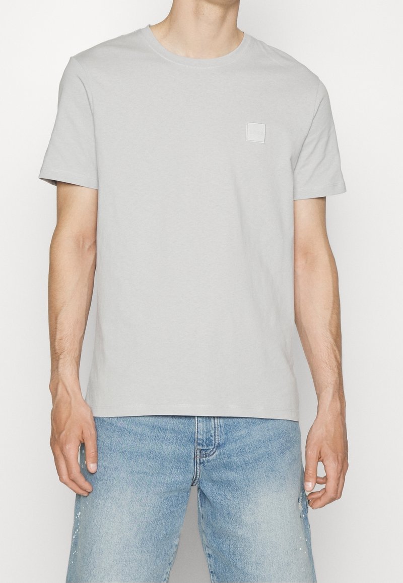 Homme portant un t-shirt gris clair uni à manches courtes et un jean bleu clair avec des éclaboussures de peinture, debout devant un fond uni.