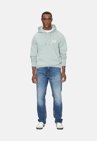 Sudadera azul claro con cordones y bolsillo frontal, combinada con jeans azules y zapatillas blancas. La sudadera presenta un pequeño detalle de logo.