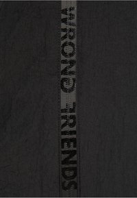 Un tessuto nero presenta una fascia verticale con le parole "WRONG" e "FRIENDS" in lettere grasse e texturizzate. Il tessuto ha una sottile texture increspata.