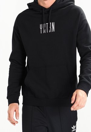 Man draagt een zwarte hoodie met witte verticale barcode-stijl tekst op de borst, gecombineerd met een zwarte Adidas broek.