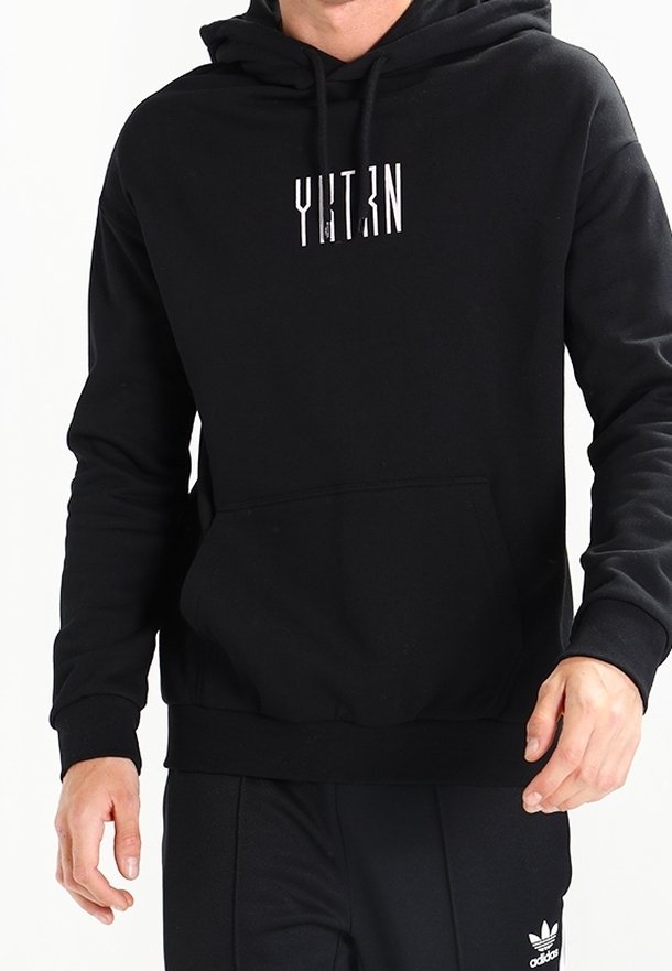 Homme portant un sweat à capuche noir avec un texte vertical blanc type code-barres sur la poitrine, associé à un pantalon noir Adidas.