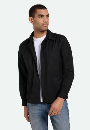 Camisa negra de botones con cuello, que incluye un bolsillo en el pecho, mangas largas y una textura suave, combinada con una camiseta blanca y vaqueros azules.