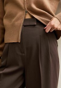 Bruine gebreide trui met een ritssluiting, gecombineerd met bruine, op maat gemaakte broek met een voorkruiszak en gladde, vallende stof.