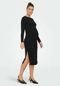 Abito premaman nero a coste con maniche lunghe, collo alto, spacchi laterali e design aderente. Indossato con scarpe nere con tacco.