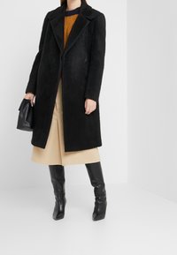 Abrigo de lana negro y texturizado con diseño de doble botonadura, gran cuello y bolsillos laterales, combinado con una falda beige y botas altas negras.