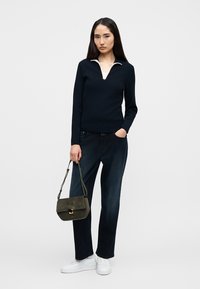 Pull bleu marine zippé avec col blanc, associé à un pantalon large en denim foncé. Le modèle tient un sac en daim vert foncé et porte des baskets blanches.