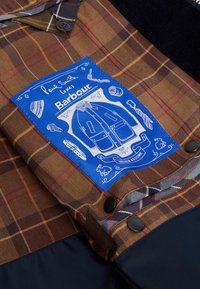 Tecido xadrez castanho com um rótulo azul contendo ilustrações e texto. Inclui botões pretos e um painel inferior em contraste.