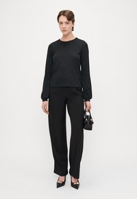 Blusa preta de mangas longas com padrão texturizado, combinada com calças pretas de cintura alta e saltos pretos de bico fino; segurando uma pequena bolsa preta.