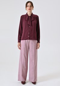 Burgunderfarbene Bluse mit gebundenem Halsband, vorn mit Knopfverschluss; kombiniert mit hellrosefarbigen, gemusterten Schlaghosen und burgunderfarbenen Schuhen.