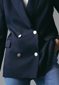 Persona che indossa un blazer doppiopetto blu navy con bottoni argentati e jeans blu, con una mano in tasca su uno sfondo grigio.