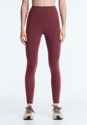 Nő sötétvörös, magasderekú leggingsben és bézs sportcipőben, egy egyszerű fehér háttér előtt állva.