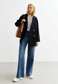 Abrigo de marina de doble botonadura, camisa a rayas, jeans de pierna ancha de mezclilla y bolsa tote marrón. Las zapatillas blancas completan el atuendo. Se aprecian tejidos texturizados.