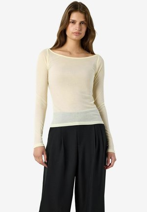 Femme portant un haut côtelé crème ajusté à manches longues et un pantalon large noir ample, debout devant un fond blanc uni.