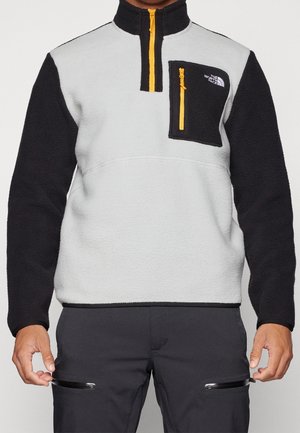 Person trägt einen zweifarbigen Fleece-Pullover mit schwarzen Ärmeln, hellgrauem Torso und gelber Reißverschlusstasche auf der Brust von The North Face.