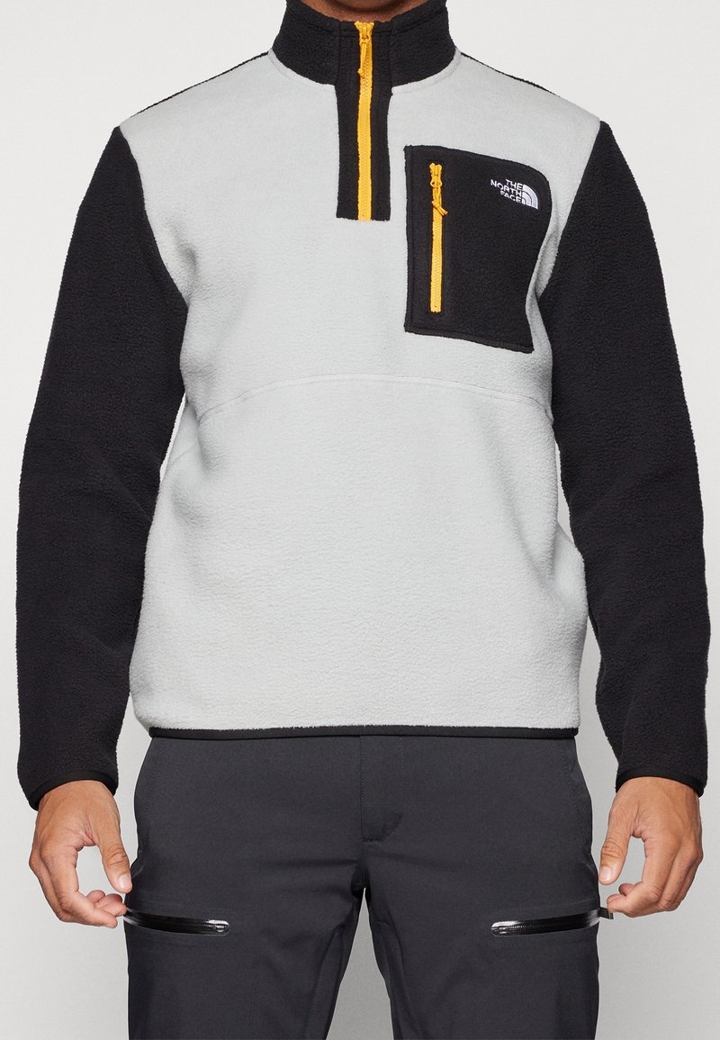 Personne portant un pull polaire bicolore avec des manches noires, un torse gris clair et une poche poitrine zippée jaune de The North Face.