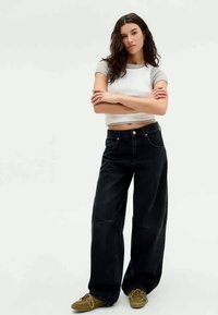 Žena stoji s prekriženim rukama, odjevena u sivi i bijeli kratki top s kratkim rukavima, visoko strukirane crne hlače širokih nogavica i smeđe cipele s vezicama.