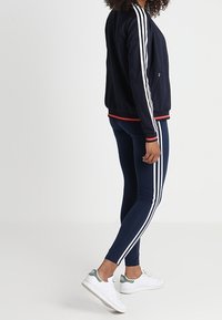 Marinblå zip-up jacka med vita ränder på ärmarna och röda detaljer, kombinerad med marinblå leggings som har vita sidostripes och vita sneakers.